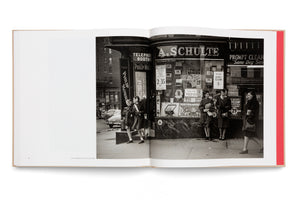 An open book showing a black-and-white photograph of a 1940s New York City street corner. Several women and two young girls in coats stand and walk on the sidewalk in front of an A. Schulte smoke shop, its window filled with advertisements for cigarettes and gum.