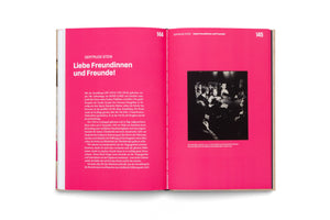 An overhead photo of an open book with bright magenta pages lying on a white surface. The book is open to pages 144 and 145, which feature German text about Gertrude Stein and a black-and-white photograph of a group of people at a gallery event.