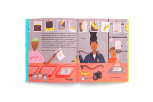 A colorful two-page spread from a German children's book illustrating the process of film photography. On the left, a person develops a photo of a banana in chemical trays. On the right, people are in a darkroom with labeled equipment like a camera, enlarger, and film. Above them, developed photos of the banana hang on a drying line.
