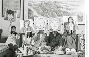 A thought-provoking black and white photograph of an Asian family of five. They are seated in a row, and each person hides their face by holding up a large, stylized portrait drawing. The drawings depict various life archetypes: a young woman, a graduate, an elderly couple, and a professional woman. The setting includes a tea set on a low table and a mountain landscape painting in the background.