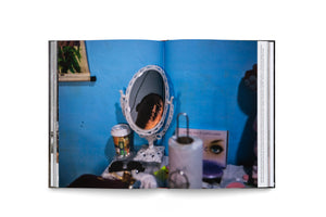 An open book on a white surface, showing a photograph of a person's reflection in a small, ornate vanity mirror. The mirror sits on a cluttered table against a bright blue wall, and the face in the reflection is half-lit, creating a somber, intimate mood.