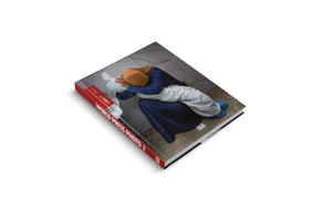 The World Press Photo 2024 book shown at an angle on a white background. The cover features a photograph of a woman in a blue dress and tan headscarf kneeling on a tiled floor, embracing a body wrapped in a white shroud. The book has a red spine with the title in white lettering.