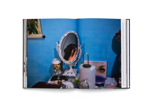 An open book laid flat on a white surface, displaying a full-page color photograph on the right. The photo shows a person's face reflected in a small, ornate, white mirror that sits on a cluttered vanity against a vibrant blue wall. The reflection is in shadow, with one side of the face highlighted by light.