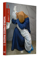 The cover of the book World Press Photo 2024. The photograph shows a woman in a blue dress and a tan headscarf, kneeling on a tiled floor. She is grieving as she embraces the body of a small child wrapped in a white shroud, holding the body close and bowing her head.