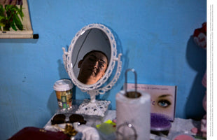 A woman’s face with a pensive expression is seen in the reflection of an ornate, white-framed oval mirror. The mirror rests on a cluttered dressing table against a bright blue wall, surrounded by everyday items like a coffee cup, sunglasses, and makeup.