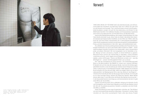 A two-page spread from an art publication. On the left, a person with short dark hair in a black jacket writes on a large black-and-white photograph of a person's bare backside, mounted on a white-tiled wall. On the right, a page of German text is titled Vorwort.