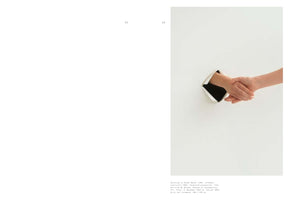A book spread showing a photograph of Yoko Ono's artwork, Painting to Shake Hands. Against a plain white wall or canvas, a square hole is cut with its corners peeled back. Two hands reach through the opening from opposite sides and clasp in a handshake.