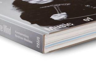 A close-up shot of the corner of a book, showing its grey spine and cover against a white background. The cover features a black-and-white photo of a person and the title Music of the Mind. The spine lists Kunstsammlung Nordrhein-Westfalen and the publisher Hatje Cantz. The book's page block is visible, revealing a light blue page among the white ones.