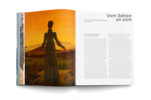 A two-page spread in an open art book. The left page features a full-page reproduction of a Romantic painting of a figure in a dark dress, seen from behind, looking out over a hilly landscape under a golden sky. The right page contains an essay in German titled Vom Sehen an sich and Zu Friedrichs Rückenfiguren.