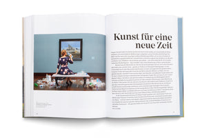 An overhead view of an open book. The left page features a photograph of a woman in a blue floral dress sitting on a bench in an art gallery. She is seen from behind, drinking from a plastic bottle while viewing a framed painting of an icy seascape. The floor around her is covered with a large pile of empty plastic bottles and containers. The right page displays German text under the large headline, Kunst für eine neue Zeit.