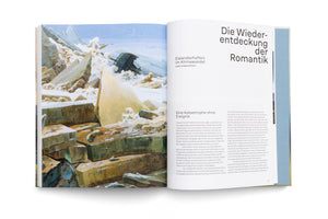 An angled, overhead shot of an open book on a white surface. The left page features Caspar David Friedrich's painting The Sea of Ice, depicting a shipwreck crushed by massive, jagged ice floes. The right page contains an article in German with the title, Die Wiederentdeckung der Romantik.