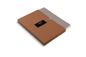 An angled, high-angle shot of a grey book titled Frequencies by Oscar Murillo, Vol. 1, partially slid out of a brown cardboard slipcase. The title is printed in white on a black label on the case. The entire object is set against a plain white background.