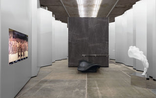 An interior view of a contemporary art gallery with concrete floors and an exposed ceiling. The space is filled with angular white partitions, a large dark marble slab with a black sculptural chair, a screen displaying a classical painting on the left wall, and a white abstract foot sculpture on a mirrored pedestal to the right.