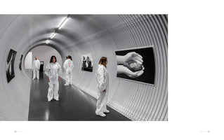 An interior shot of an art gallery in a long, tunnel-like corridor with corrugated metal walls. Four people wearing white protective coveralls are looking at black and white photographs displayed along the curved walls, one of which shows a pair of hands holding a small bird.
