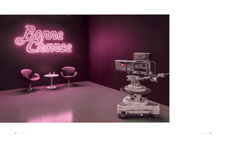 A television studio set for an interview, bathed in a magenta glow. In the foreground, a large professional broadcast camera is pointed towards two empty, modern purple chairs and a small table. On the dark wall behind them, a bright pink neon sign in cursive script reads Bonne Chance.