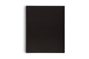 A minimalist, top-down photograph of a closed, black, hardcover book with a plain cover, positioned vertically on a white background. A soft shadow is visible along its left edge, and the spine is on the right.