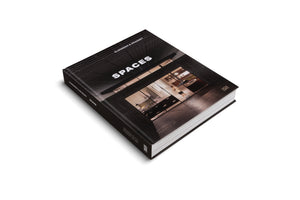 A studio photo of a thick, black hardcover book titled SPACES by Elmgreen & Dragset, angled on a white surface. The cover features a photograph of a modern interior viewed from above through a dark, grid-like ceiling structure.