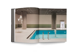 An open book displays a two-page photograph of a sterile, minimalist indoor swimming pool with large concrete pillars. The surreal scene contains three small, all-white figures: one sits on a high lifeguard chair with binoculars, another in a protective suit stands by the water's edge, and a third looks out a door.