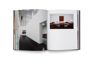 An open book on a white background displays a two-page photo spread. On the left, a person hangs from the high ceiling of a modern, white industrial-style building. On the right, a person in pink pajamas lies on their side across a long conference table, with a large pink plush toy sitting in a chair at the end.