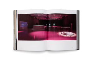 An open book displays a two-page photograph of a spacious, futuristic room bathed in pink light. On the polished wooden floor in the foreground, a small white kitten sits on a spotlit, circular platform with a target design. In the background, there is a curved white bar and several glowing, circular targets mounted on the far wall.