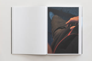 A high-angle shot of an open book on a white surface. The left page is blank, while the right page features a full-bleed color photograph taken from a first-person perspective, looking down at the photographer's own legs in dark jeans, with a hand resting on their thigh.