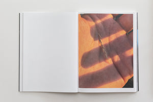 An open book lies flat on a white surface. The left page is blank, while the right page displays a close-up photograph of a hand holding a dry sprig of grass. The hand is bathed in warm, golden light, with dark, linear shadows cast across the palm.