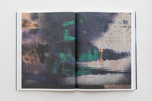 An overhead view of an open book on a white surface, displaying a two-page spread of an abstract image. The image features a textured, weathered surface with layers of dark purple, black, and green paint, alongside areas of distressed off-white and a small patch of orange.