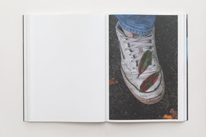 An open book laid flat on a white surface. The left page is blank, and the right page shows a full-page color photograph of a dirty white sneaker with blue jeans, seen from above. Two small, pointed leaves, colored red and green, rest on the shoe.