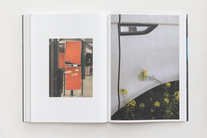 An overhead view of an open book on a white surface. The left page displays a small photograph of a subway station with orange, gridded signs. The right page features a large, close-up photograph of the side of a white car, with yellow wildflowers and a white butterfly next to the wheel well.