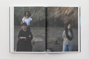 An open book laid flat, showing a two-page color photograph of three women on a sandy beach in front of a large rock cliff. On the left page, a woman in black with arms crossed is in the foreground, with another woman in a white shirt behind her. On the right page, a young woman in a striped shirt and jeans stands with her hands behind her back. The three women look in different directions with neutral, pensive expressions.