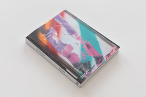 An overhead, angled shot of a book titled Remnants by Paul Hutchinson, lying on a white surface. The book's cover features an abstract, colorful design with what appears to be spray paint strokes in purple, pink, teal, and orange. The subtitle reads Selected Works 2019-2024.