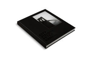 A product shot of a black hardcover book, City Visions by Peter Arnell, lying at an angle on a white surface. The matte black cover features a black and white photograph of a suspension bridge tower partially obscured by fog, with the title in a glossy black font below it.