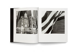 An open book with a black cover displays a two-page spread of black and white photographs on a white surface. The left page shows a brick church on a wet city street, dwarfed by modern skyscrapers. The right page shows a close-up, angled view of an American flag's stripes next to a corrugated metal surface.