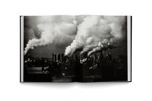 An open book, seen from above on a white background, displaying a two-page, grainy black-and-white photograph. The photo shows a silhouetted city skyline with massive plumes of white smoke rising from numerous buildings and filling the sky. A body of water is in the foreground.