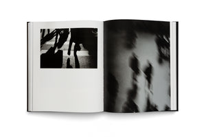 An open, black-covered book is shown on a white background. The left page features a stark, black and white street photograph of legs and long shadows crossing a sunny crosswalk. The right page displays a full-bleed, grainy, and blurry black and white abstract photograph with a sense of motion.
