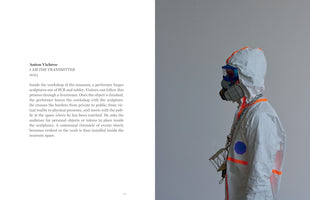 A two-page spread from a book. On the left, black text describes the art piece I AM THE TRANSMITTER by Anton Vichrov. On the right, a photograph shows a performer in profile wearing a white hazmat suit with orange stripes, a respirator mask, and goggles against a gray wall. Heavy chains around their neck hold a small, cage-like metal sculpture.