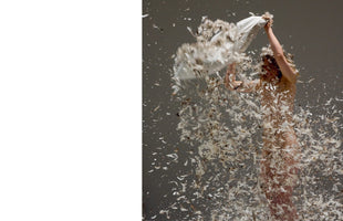 A nude person stands on the right side of the image against a gray background, vigorously shaking a white pillow and releasing a chaotic storm of feathers that swirls around their body. The left side of the image is a solid white panel.