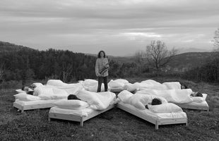 A surreal black and white photograph of a person with long curly hair holding a saxophone, standing in a grassy field. They are surrounded by multiple beds arranged in a radial pattern, each occupied by people sleeping under white duvets. In the background are forested hills and distant mountains.