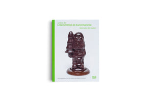 The front cover of the German book Lexikon der Lebensmittel als Kunstmaterial, which translates to Encyclopedia of Food as Art Material. The cover features a dark chocolate sculpture of Santa Claus, whose pointed hat has been replaced with a shape resembling a butt plug. The book has a bright green spine and is set against a plain white background.