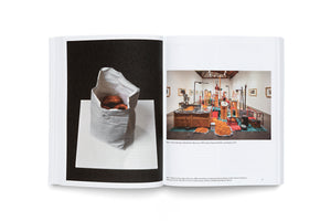 An open art book is displayed against a white background. The left page shows a photograph of a white paper bag with donuts inside against a black backdrop. The right page shows a photograph of a large, complex art installation in a gallery, featuring machinery and piles of orange, donut-shaped objects.