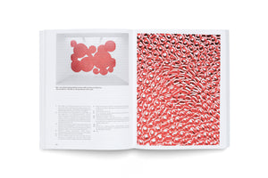 A top-down view of an open art book. The left page shows German text and a small photo of a red sculpture made of circles. The right page is a full-page close-up, revealing the sculpture is made of a dense texture of red and white gummy candy vampire teeth.