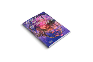 A three-quarter view of the paperback book Quantum Soup by Libby Heaney, published by Hatje Cantz, resting on a white surface. The cover features a vibrant, surreal illustration of a large pink, translucent, jellyfish-like form entangled in dark, thorny branches against a purple and blue background.