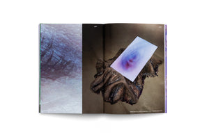 An overhead shot of an open art book on a white surface. The left page is a full-bleed abstract image with blue and purple textures. The right page shows a photograph of an installation: a rectangular screen with a blurry, colorful image rests on a dark, crinkled sculpture on a concrete floor. The pages are numbered 122 and 123.