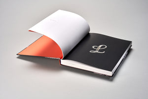 A black hardcover book rests open on a light grey surface. A white page is partially turned, revealing a vibrant orange endpaper. The facing page is solid black with an ornate, silver, cursive letter L embossed in the center.
