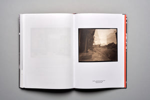 An overhead shot of an open book on a gray surface. The left page is mostly white with a faint, faded print. The right page displays a square, sepia-toned pinhole photograph of a street scene with a caption printed underneath.