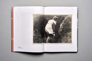 An overhead shot of an open book on a gray surface. The book displays a two-page spread, with a vintage black-and-white photograph on the right page. The photo shows a woman in a summer dress, seen from behind, bending over in a lush, sunlit garden. The left page is mostly blank, with a small caption at the bottom.
