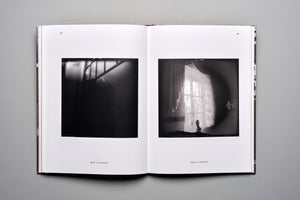 An overhead shot of an open photobook on a gray surface. The two-page spread displays two grainy, black-and-white photographs. The left photo is a dark interior showing the shadow of a railing. The right photo shows a blurry, sunlit window with sheer curtains and a small silhouette on the sill.