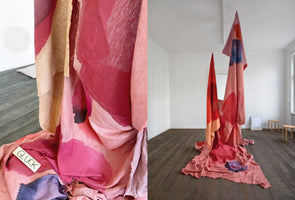 A diptych showing a textile art installation in a gallery. The image on the right shows two long, abstractly dyed fabrics in shades of red, pink, and purple hanging from the ceiling and pooling on the dark wood floor. The image on the left is a close-up of the draped fabric, showing a small wooden tag with the word GLÜCK on it.