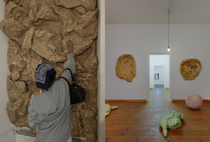 A diptych showing an art gallery. On the left, a person wearing a grey hoodie and a blue bandana touches a large wall installation of crumpled brown paper decorated with colorful dots and white drips. On the right, a wider view of the gallery shows abstract sculptures on the white walls and soft, stuffed forms on the wooden floor.