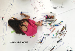 An overhead view of a young girl in a pink outfit, lying on a large sheet of white paper on the floor. She is drawing on a life-sized body outline, which is part of a larger art project filled with other drawings and handwritten text. Many colorful markers and crayons are scattered around her. The text WHO ARE YOU? is written at the bottom of the image.